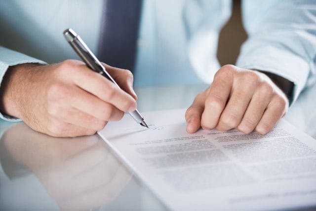 A close-up of an elder law attorney signing a contract. He is wearing a dress shirt and tie.