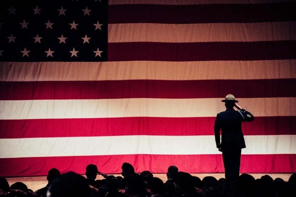 A silhouette of man in inform faces aways from the camera and salutes the American flag on a stage.