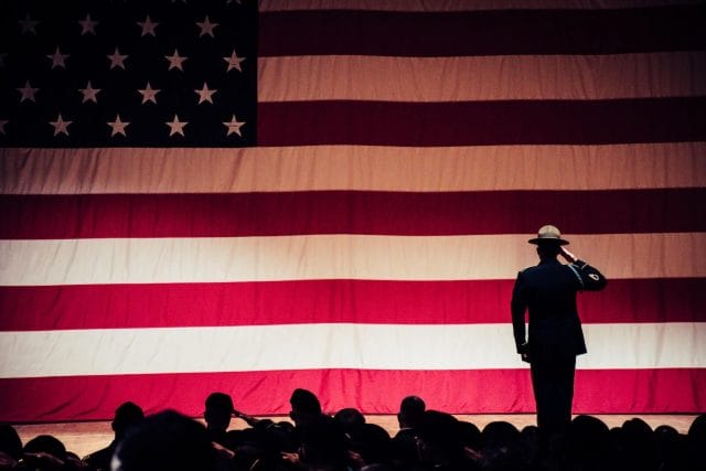 A silhouette of man in inform faces aways from the camera and salutes the American flag on a stage.