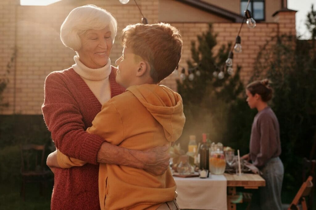 An older adult woman smiles and hugs her grandchild during an outdoor family gathering.