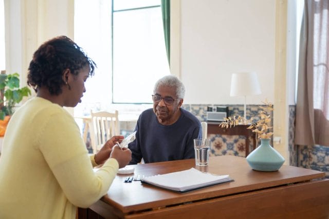 A senior man sits at a table with a younger caregiver as she looks at a receipt.