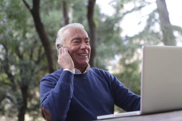 An older adult man sits in front of a laptop computer and smiles while he talks on a cell phone.
