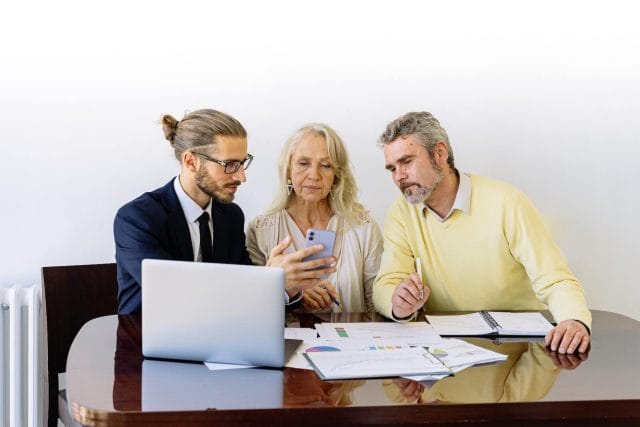 A young business man holds a phone and an older couple looks at it. There are financial documents on a desk in front of them.