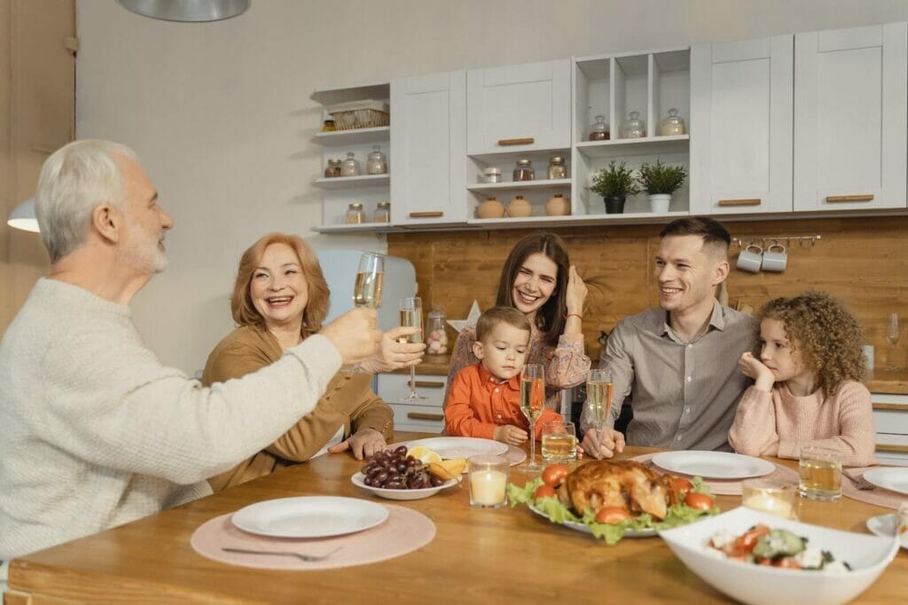 A multi-generational family sits around a table for a meal. They smile and the grandfather makes a toast.