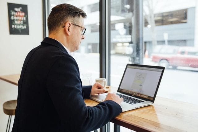 A professional man works on a laptop computer at a counter in a cafe that faces the window.