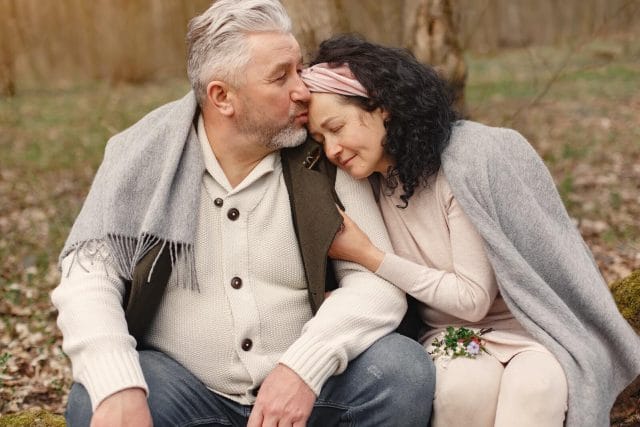 An older adult couple hugs as they sit outside on a cool fall day.