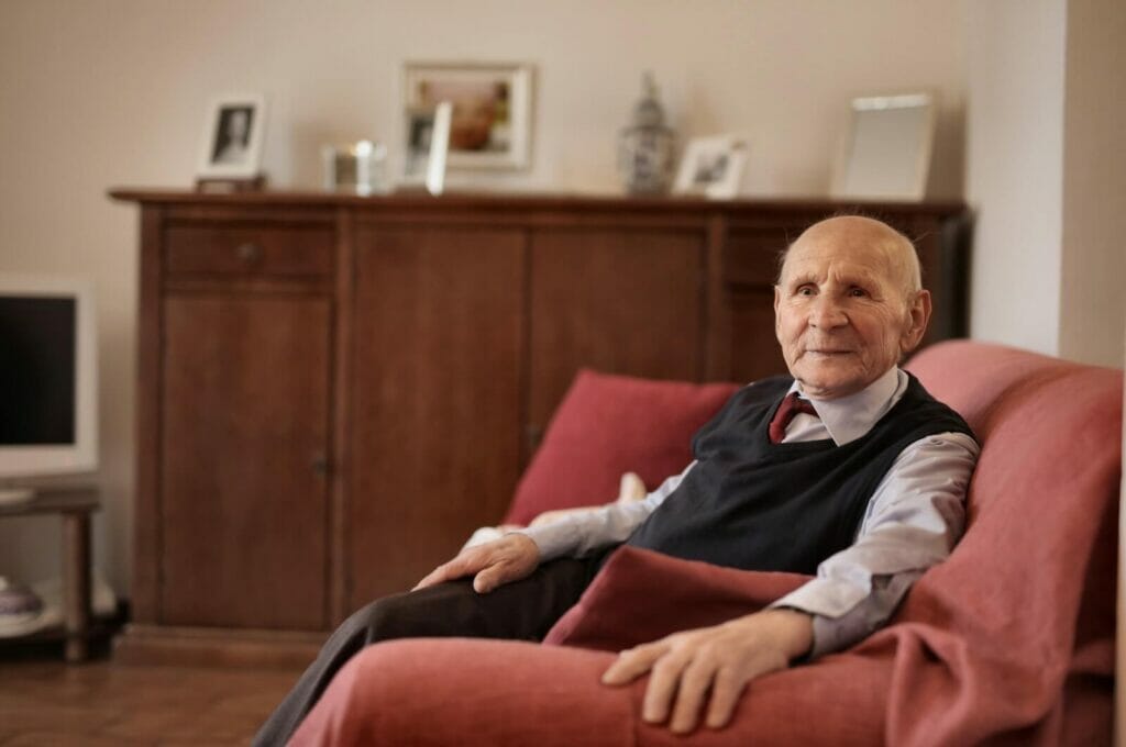 An older adult man sits on a couch and turns to smile at the camera.