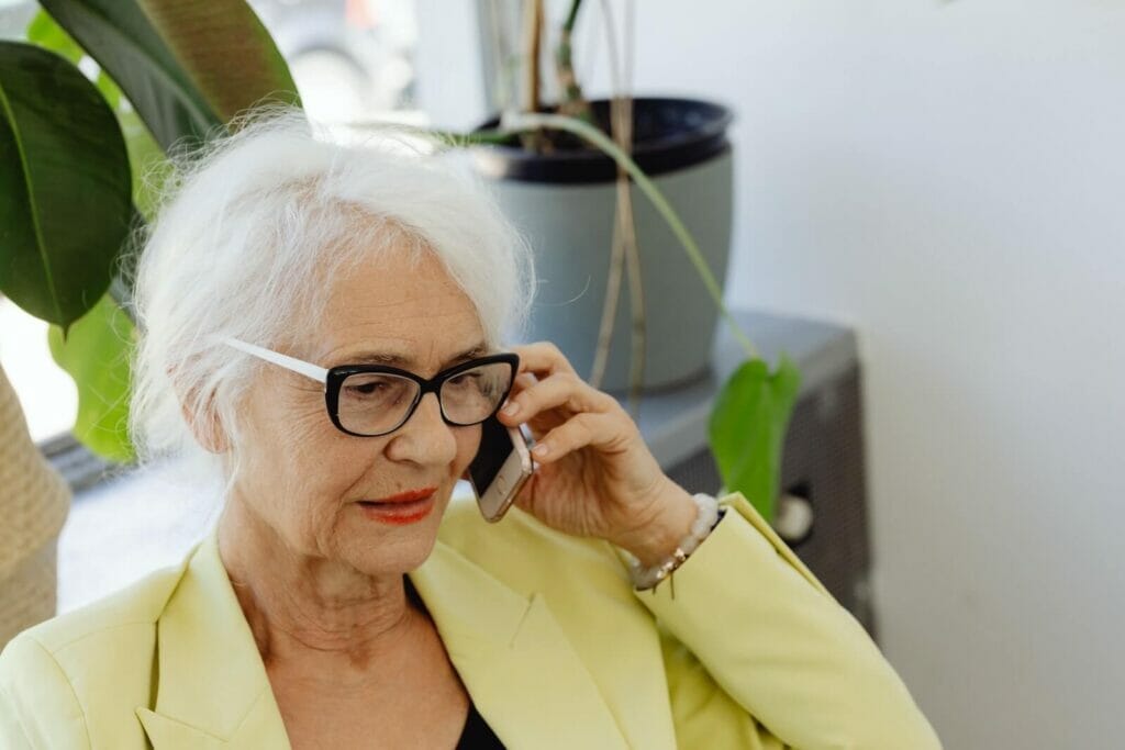 An older adult woman wears a blazer and glasses and talks on a smart phone.