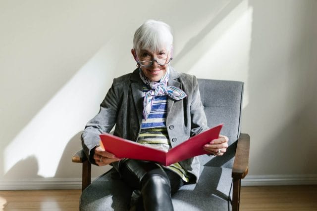 An senior woman is dressed in modern business clothing. She looks up from a file folder with documents to smile at the camera.