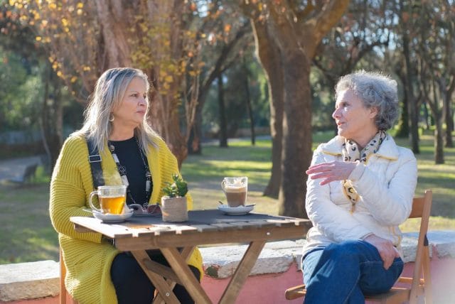 Two senior women sit at a cafe table outside with tea and coffee and have a discussion.