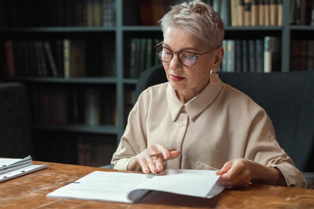 A senior woman sits at a desk in an office and reviews a contract before signing it.