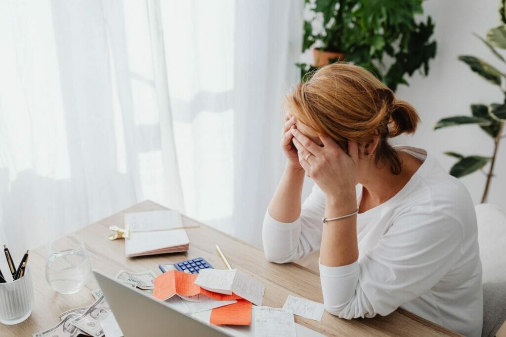 A distressed woman sits in her home office with a pile of receipts, notebook, and calculator.