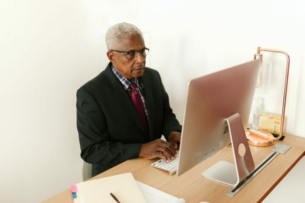 A senior man sits at a desktop computer and types.