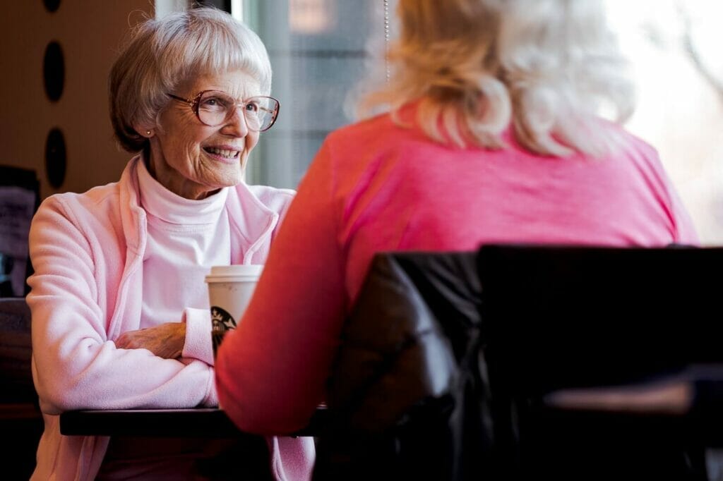 Two older adult women sit in a coffee shop. One woman smiles at the other.