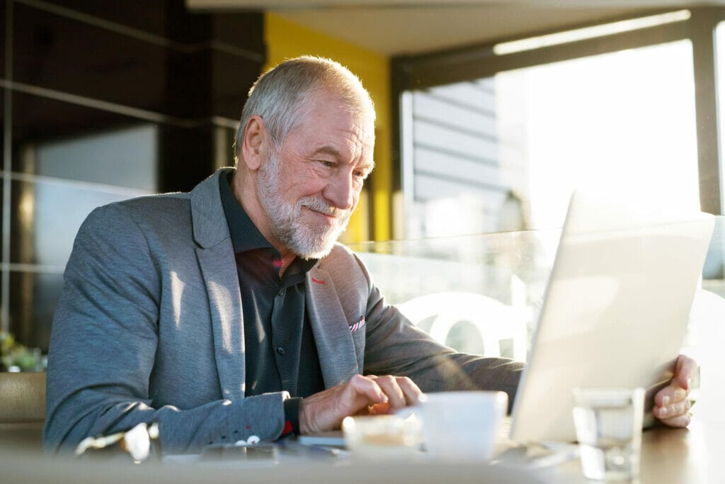 An older adult man sits in a well-lit office. He works on a laptop computer.