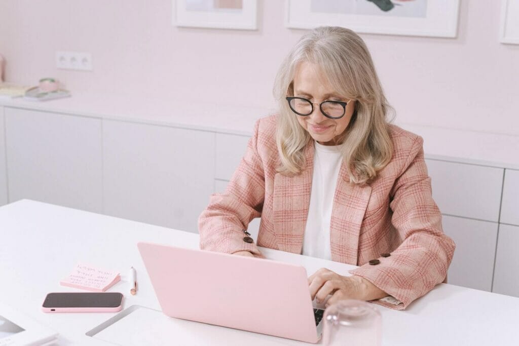 An older professional woman sits at a desk and types on a laptop computer.