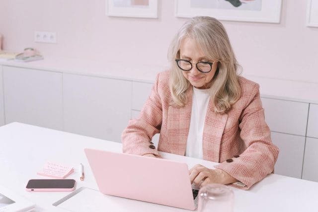 An older professional woman sits at a desk and types on a laptop computer.