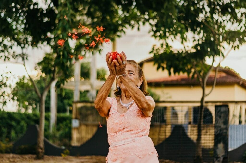 An older adult woman wears a dress. She appears happy as she tosses flower petals in the air.