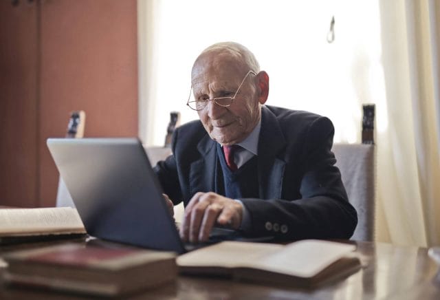 An older adult man wearing a suit and reading glasses types on a laptop computer while seated at a desk.