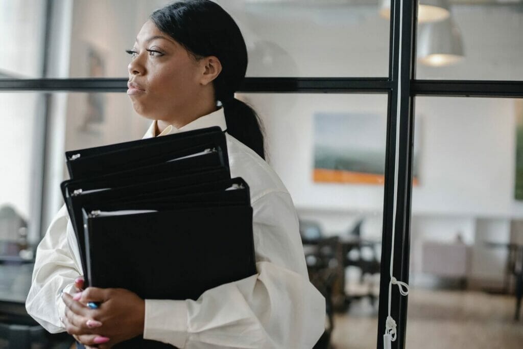 A young business woman holds binders of information as she stands in front of an interior office window.