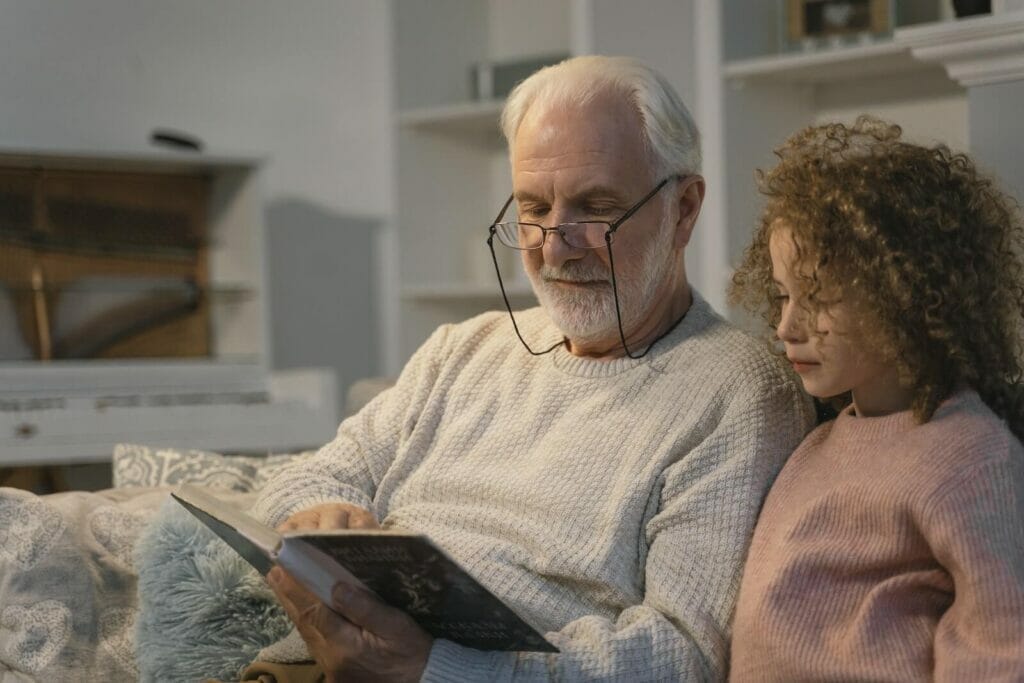 A senior man wearing glasses reads a book to a young girl.