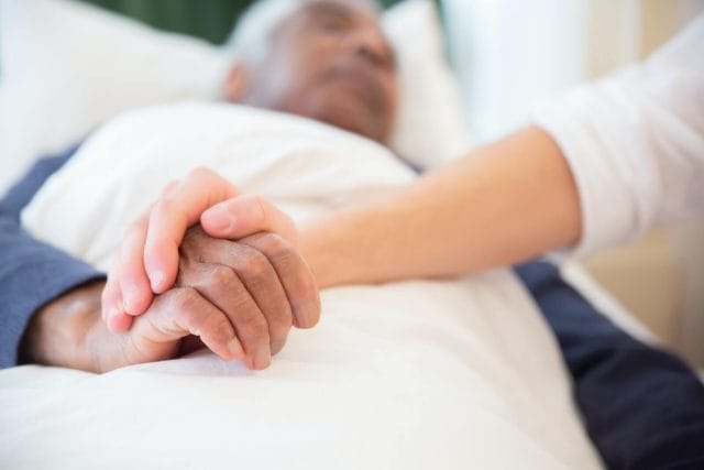 An image of a senior man laying in bed. A caregiver lays their hand over his and holds it.