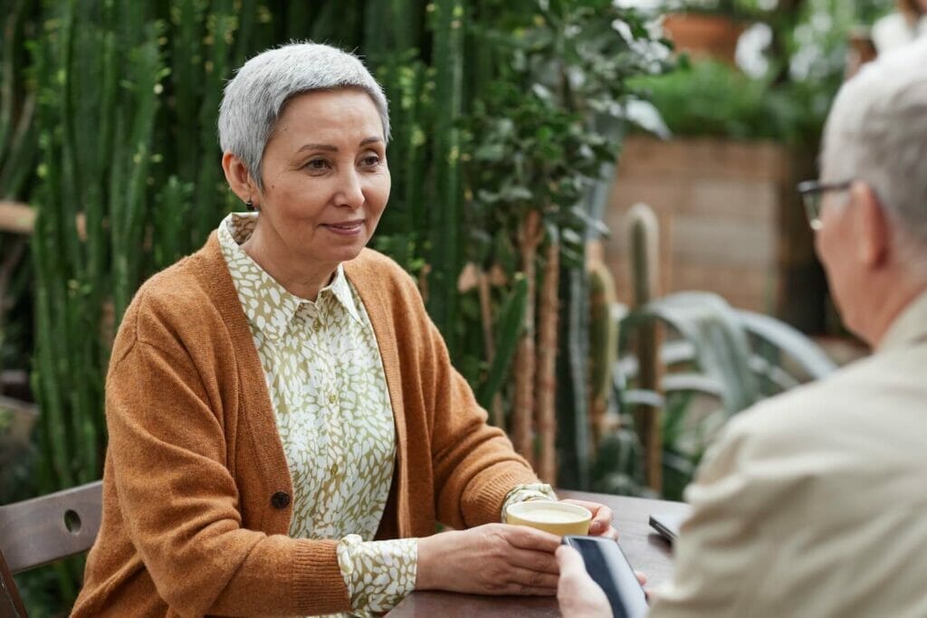 A senior woman sits across from a friend at a table. She holds a coffee mug and smiles at him.