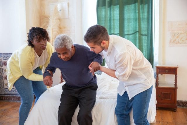Two young caregivers help an older adult man stand up.