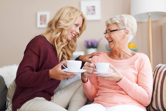 A youthful woman and senior woman smile at each other as they hold coffee cups and sit on a couch together.