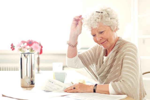 A senior woman sits at a table in a brightly lit room of her home. She reads a newspaper.