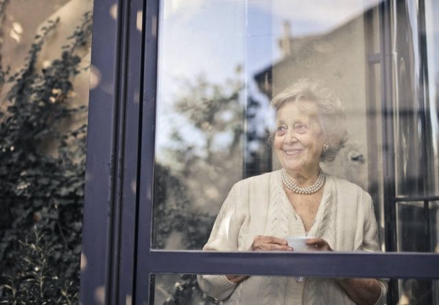 An older adult woman stands inside her home, looking out of a large window, holding a tea cup.
