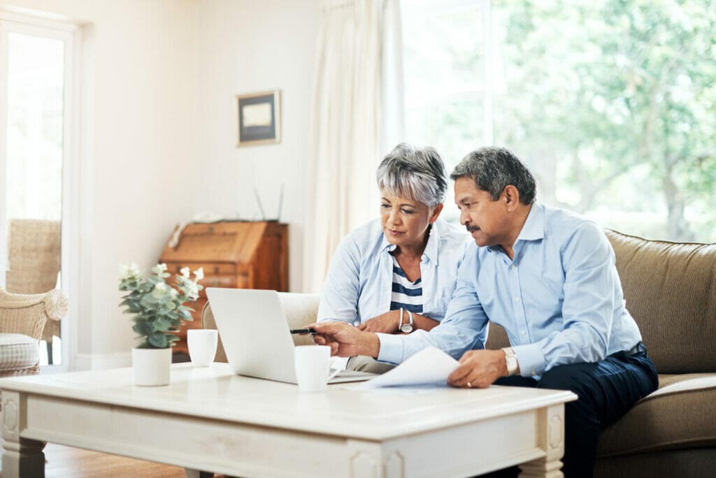 An older adult couple analyzes information on a laptop computer screen. They sit on a couch in a living room with coffee cups beside them.