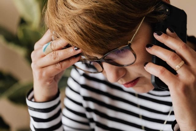 A concerned woman holds her head in her hand while she makes a phone call on a smartphone.