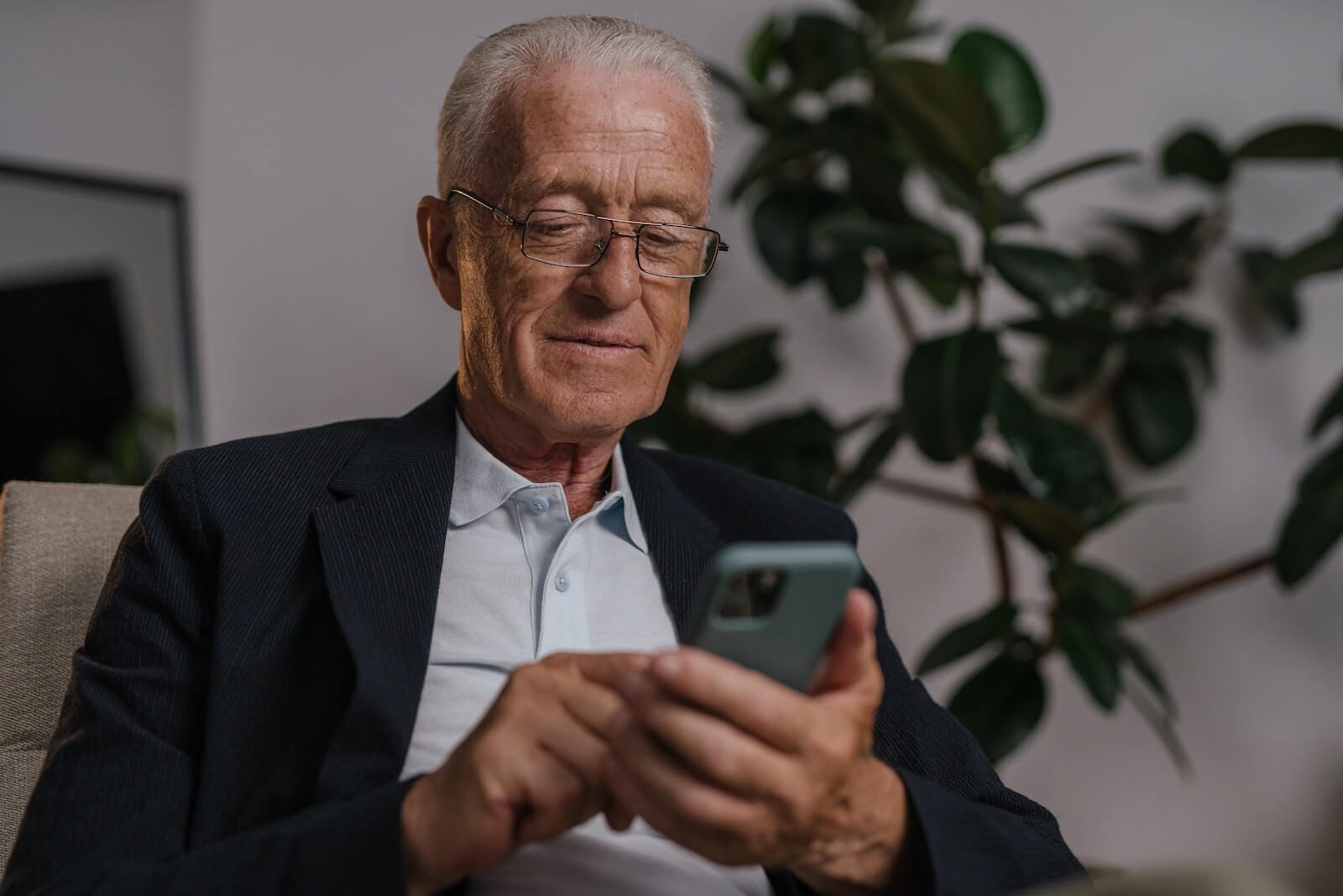 An older adult man wearing casual business clothing and glasses looks down at his smartphone.