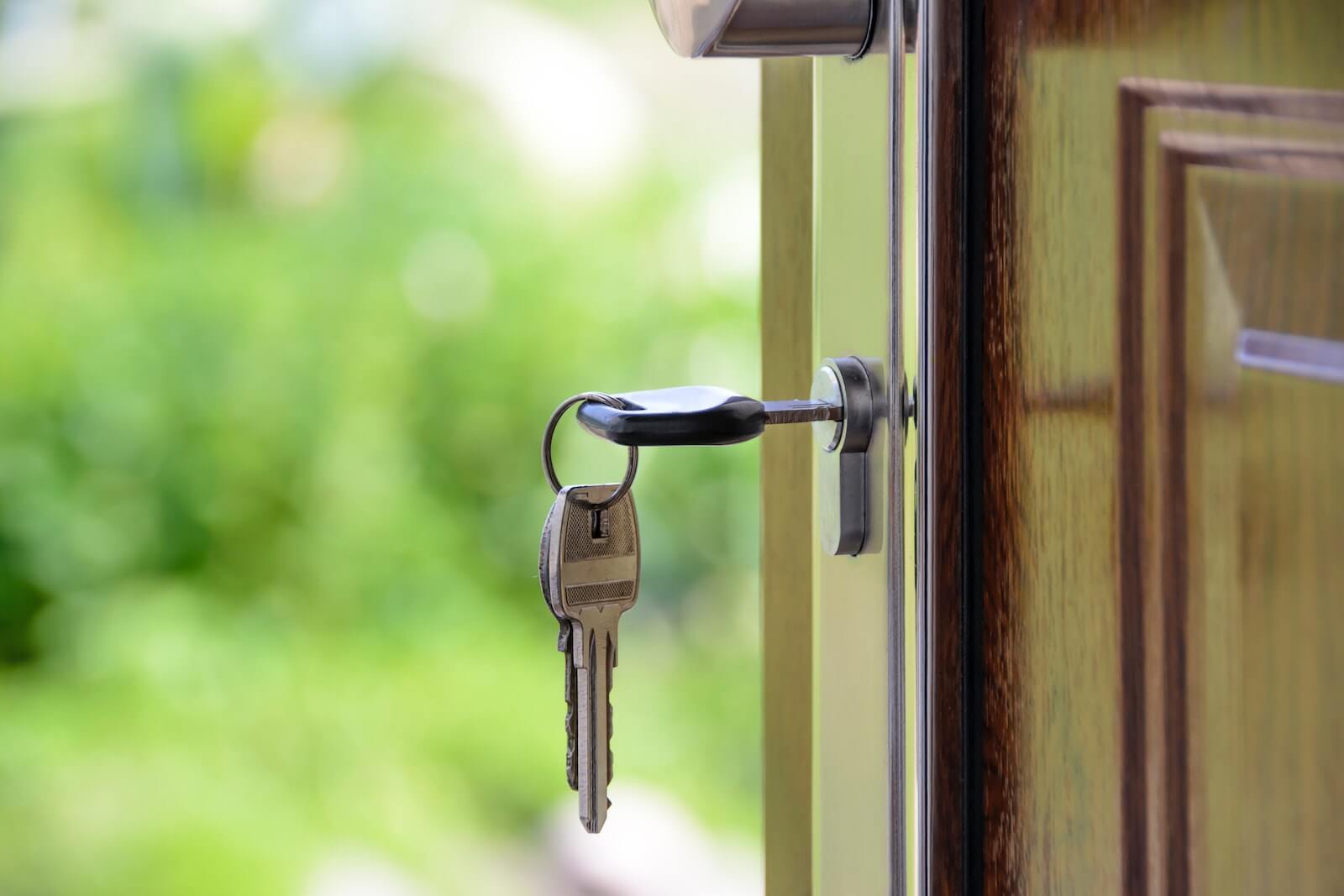 A close-up image of a key in the lock of a home's front door. There are two other keys on the key chain, and greenery is out of focus in the background.