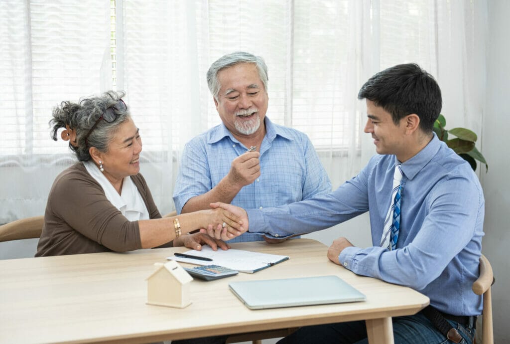 And older adult couple closes a loan with a financial professional. The woman shakes the professional's hand. The man next to her smiles.
