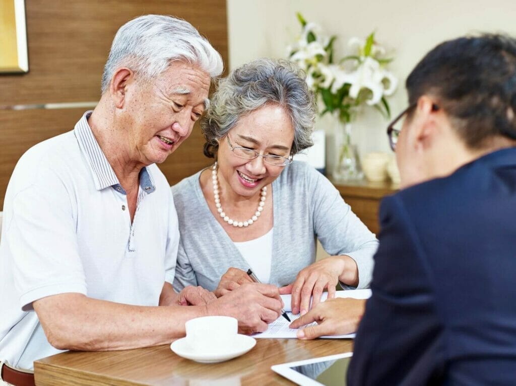 An older adult couple sits at a table with a younger man, doing some paperwork. The younger man points to a spot on the paper, and the older man is signing it. The older woman smiles as she reads along.