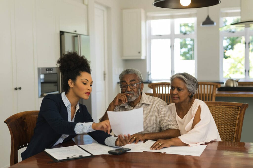 A real estate agent sits at the dining room table in a home with an older adult couple. She points to a part of a contract and the couple looks at the paper.