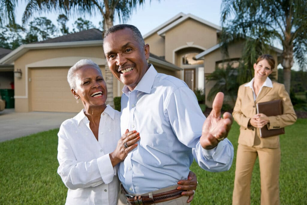 A mature couple smiles happily outside a home they sold. The real estate agent stands in the background.