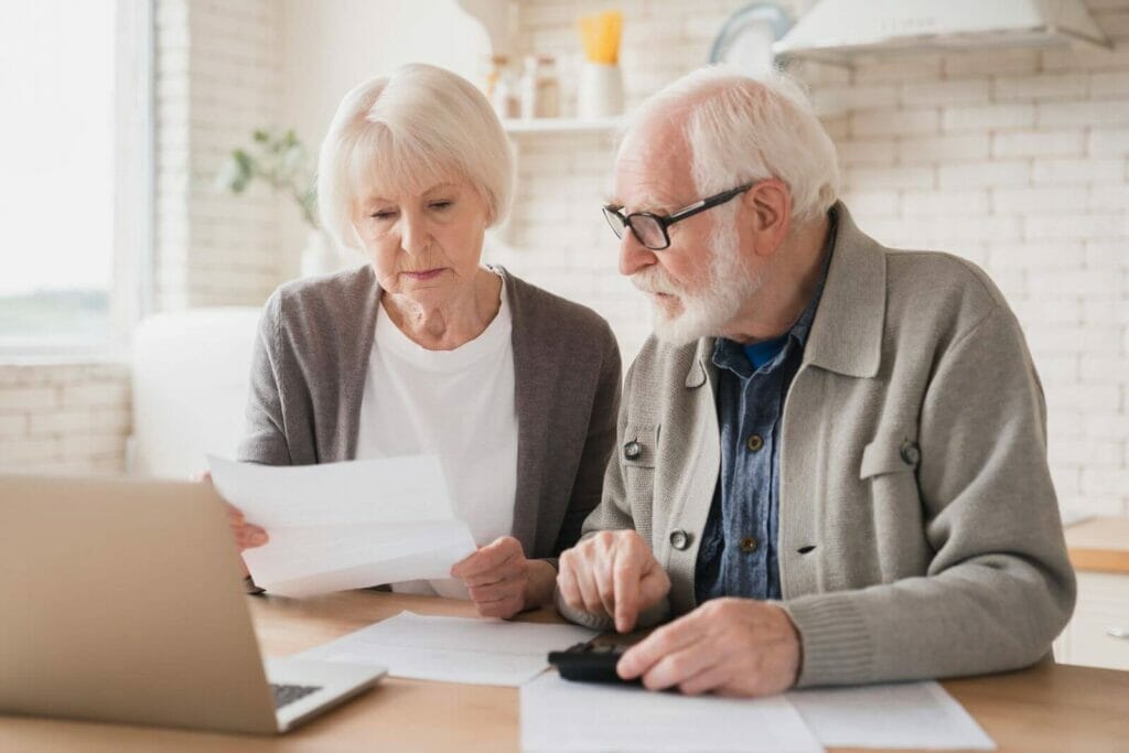 An older adult man and woman sit at a table looking over paperwork in front of a laptop. The man is holding a calculator.