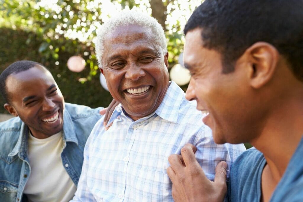 An older adult man sits between two younger men. They all smile.