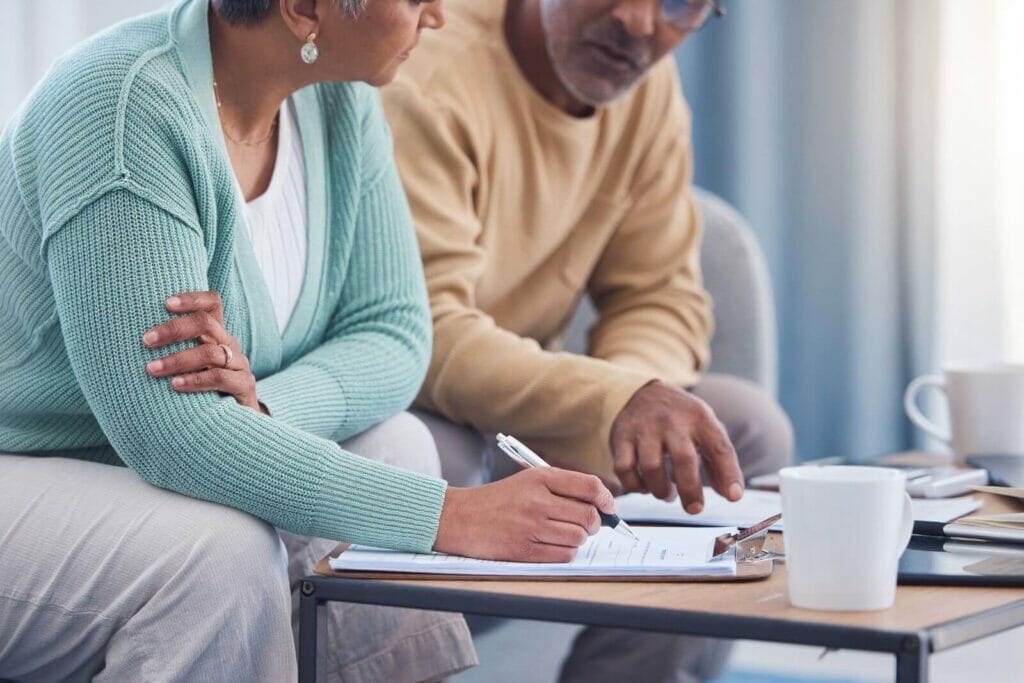 An older adult couple sits on a couch looking over paperwork on a coffee table.