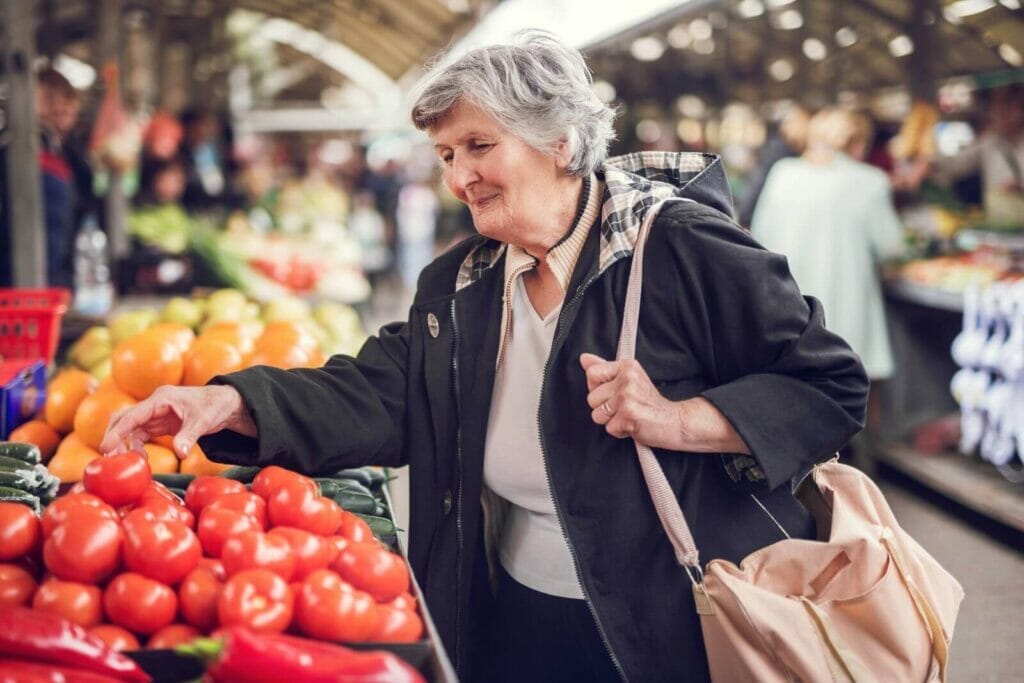 An older adult woman reaches for a tomato at a farmers market.