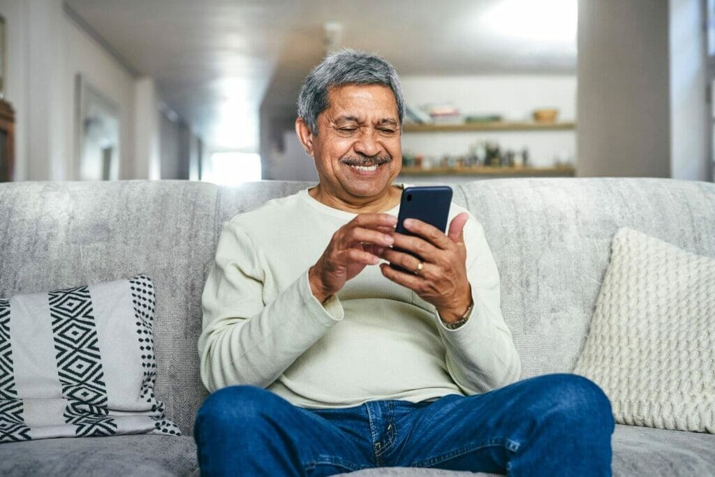 An older adult man sits on a couch, holding a cell phone and smiling.