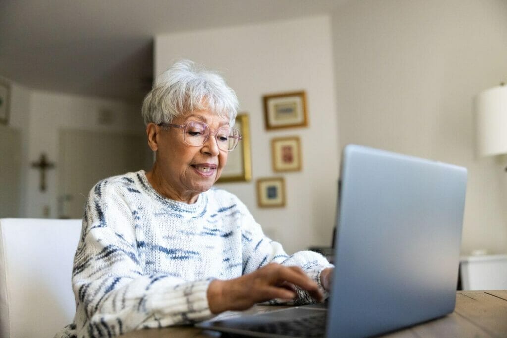 An older adult woman sits at a table using a laptop computer.