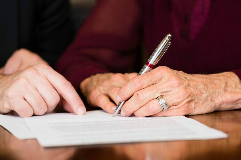 An older adult woman's hands sign paperwork where a man is pointing.