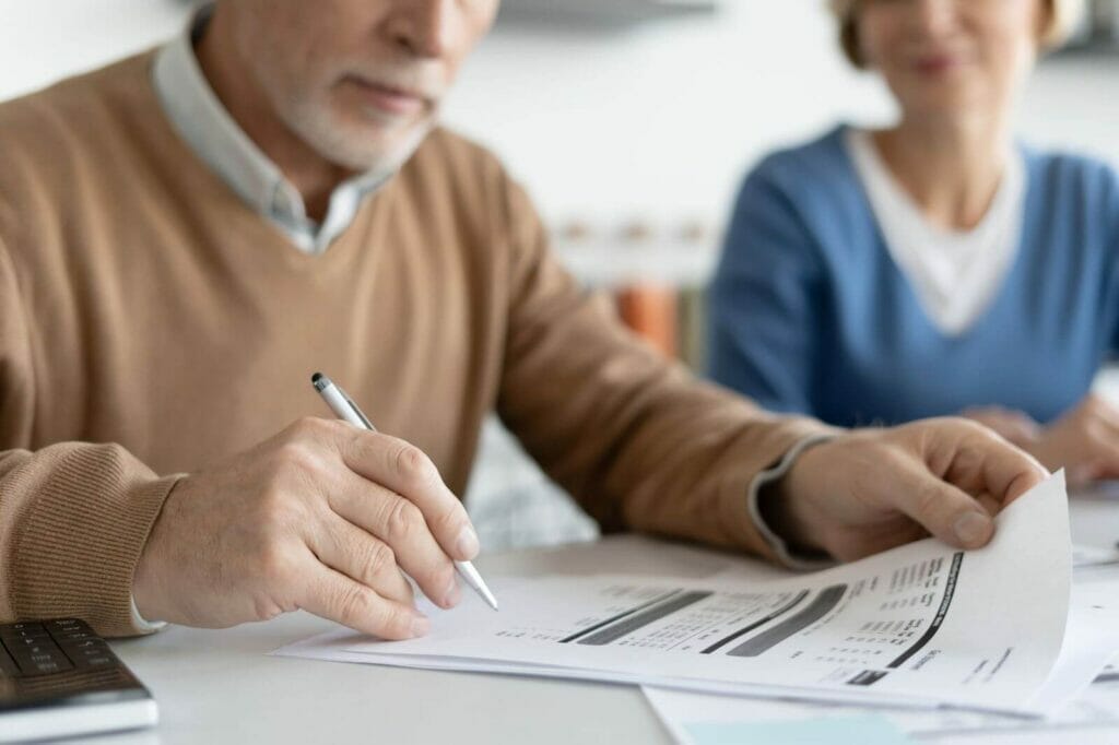 An older adult man sitting at a table holds a pen and looks at some paperwork while an older adult woman looks on.