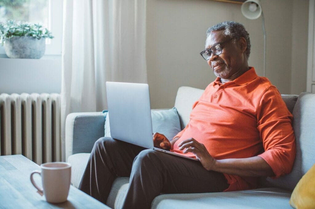 An older adult man sits on a couch using a laptop.