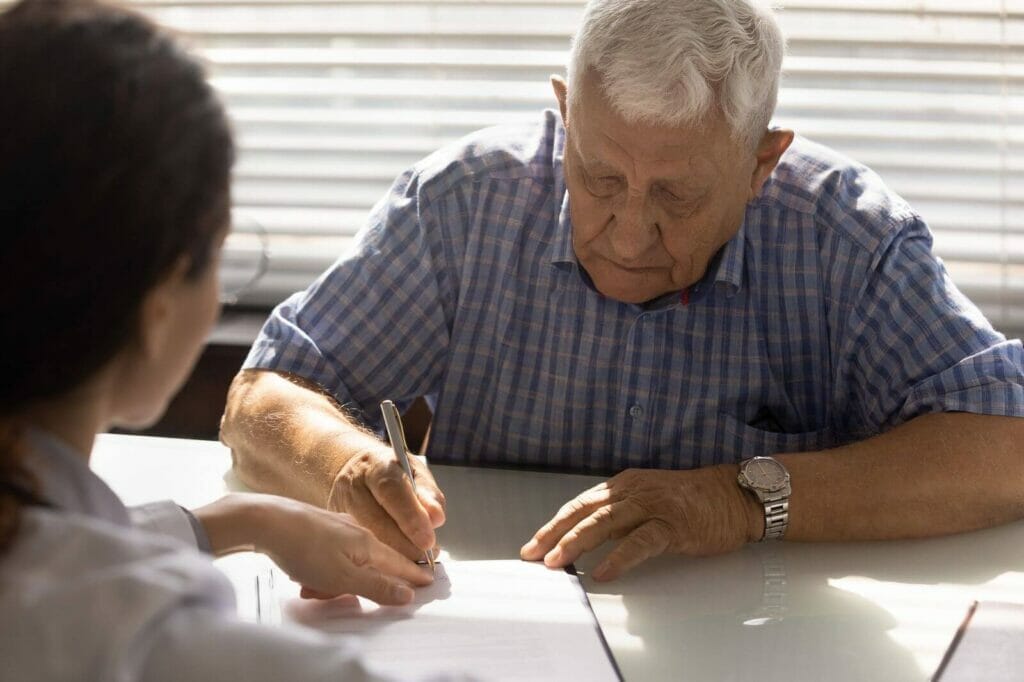 An older adult man signs a document where a younger person indicates.