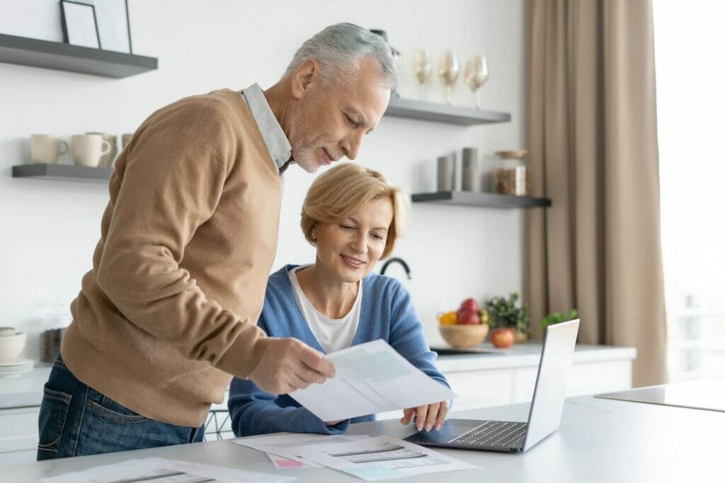 An older adult woman sits and an older adult man stands in front of a counter. There is a laptop in front of the woman, and the man is holding paperwork.